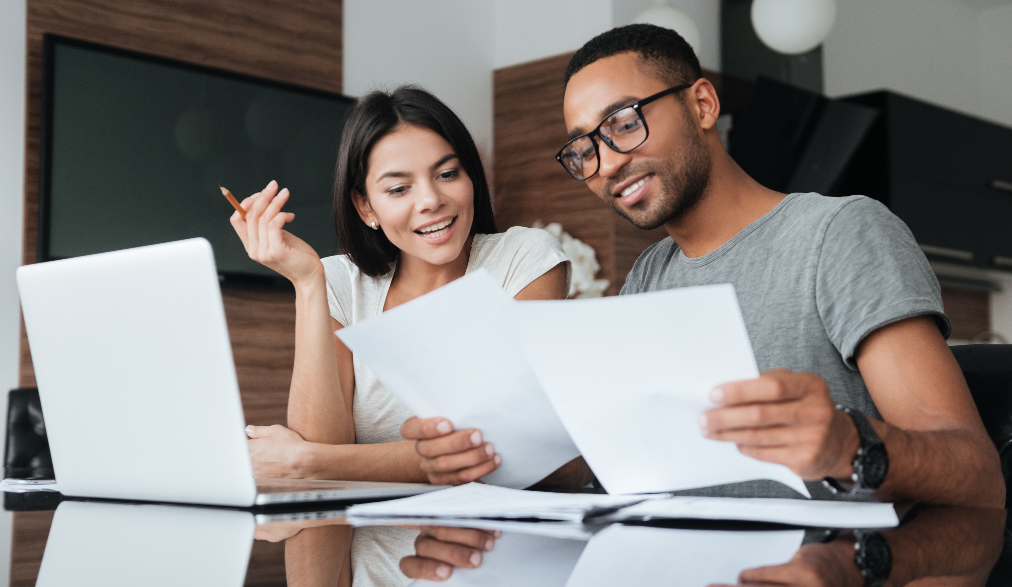 A smiling couple sits at a table reviewing documents together. The man, wearing glasses and a gray shirt, holds a sheet of paper while the woman, holding a pencil, looks at the papers with interest. An open laptop sits in front of them, and they appear to be working in a modern home setting.